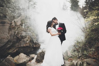 Birbirlerine aşık düğün çifti güzel manzarada kayaların yanında öpüşüyor ve sarılıyorlar.