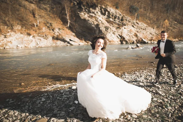 Elegante elegante novia morena feliz y hermoso novio en el fondo de un hermoso río en las montañas —  Fotos de Stock