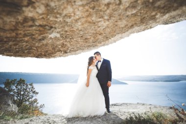 Düğün çifti mavi denizin yanındaki kayalarda öpüşüyor ve sarılıyor.