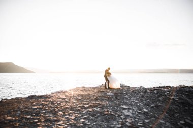 Düğün çifti mavi denizin yanındaki kayalarda öpüşüyor ve sarılıyor.