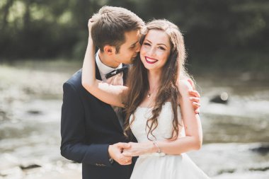 Güzel düğün çifti bir dağ nehrinin kıyısında taşlarla öpüşüp kucaklaşıyorlar.