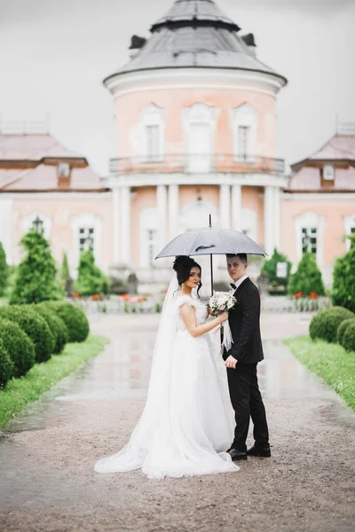 雨が降っている間、美しい若い結婚式のカップルは傘の下で手の花の花束でポーズ — ストック写真