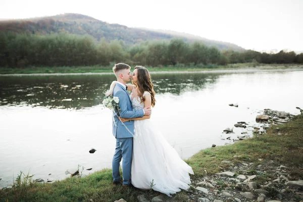 Elegante novia rubia feliz y elegante novio en el fondo de un hermoso río en las montañas —  Fotos de Stock