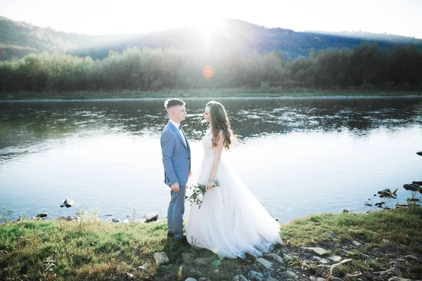 Elegante novia rubia feliz y elegante novio en el fondo de un hermoso río en las montañas — Foto de Stock