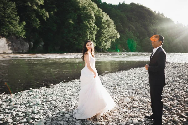 Elegante novia rubia feliz y elegante novio en el fondo de un hermoso río en las montañas —  Fotos de Stock