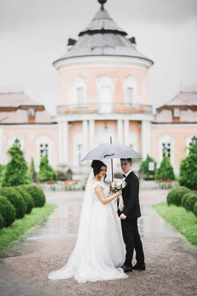 雨が降っている間、美しい若い結婚式のカップルは傘の下で手の花の花束でポーズ — ストック写真