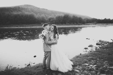 Zarif, şık, mutlu, sarışın gelin ve dağlardaki güzel bir nehrin arka planında harika bir damat.