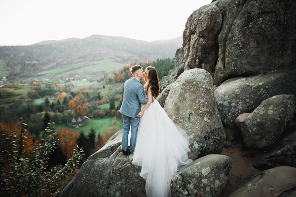 美しい豪華な花嫁は新郎にポーズをとって、素晴らしい景色、テキストのためのスペース、結婚式のカップルで山の近くで楽しんでいます — ストック写真