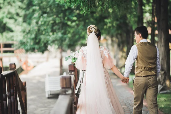 Elegante coppia di sposi felici che passeggiano nel parco il giorno del loro matrimonio con bouquet — Foto Stock