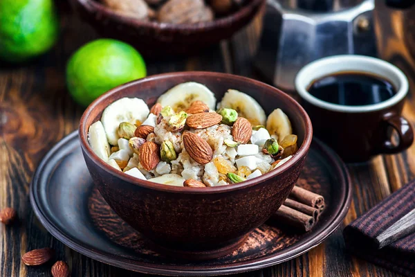 Gachas de avena ecológica con plátanos, miel, almendras, pistacho, coco, kiwi, canela, pasas en tazón de cerámica oscura con taza de café. Desayuno saludable y concepto de dieta en mesa de madera . — Foto de Stock