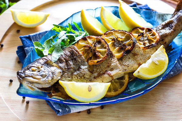 Hot fresh savory grilled whole trout barbeque with fresh herbs and lemon, served black pepper and arugula salad leaves on blue plate wooden background top view close up