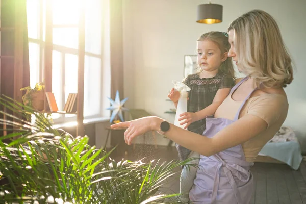 Jonge aantrekkelijke vrouw met haar peuter meisje water geven planten in huis. Huishoudelijk werk — Stockfoto