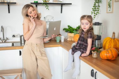 Genç anne, küçük kızıyla birlikte evde telefon ve dizüstü bilgisayarla çalışıyor.