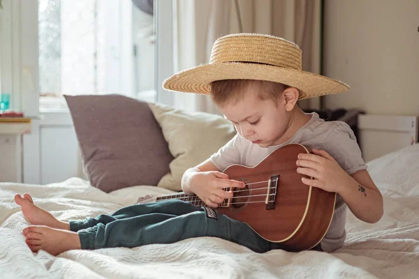 Kleine peuter jongen met hoed spelen ukelele gitaar thuis, rustieke stijl. Levensstijl concept — Stockfoto