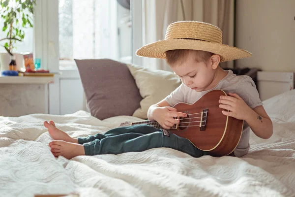 Kleine peuter jongen met hoed spelen ukelele gitaar thuis, rustieke stijl. Levensstijl concept — Stockfoto