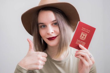 İsrail 'li genç bir kadın beyaz arka planda seyahat pasaportu taşıyor. Büyük gülümsemeyle mutlu. Tamam işaret, parmaklarla baş parmak, mükemmel işaret.
