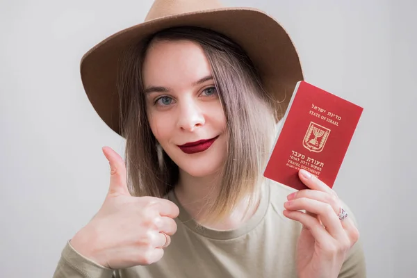 İsrail 'li genç bir kadın beyaz arka planda seyahat pasaportu taşıyor. Büyük gülümsemeyle mutlu. Tamam işaret, parmaklarla baş parmak, mükemmel işaret.