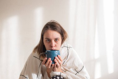 Beyaz arka planda çay içen, beyaz battaniye altında hasta bir kadın. Hastalık tedavisi, evde tecrit, karantina. Boşluğu kopyala