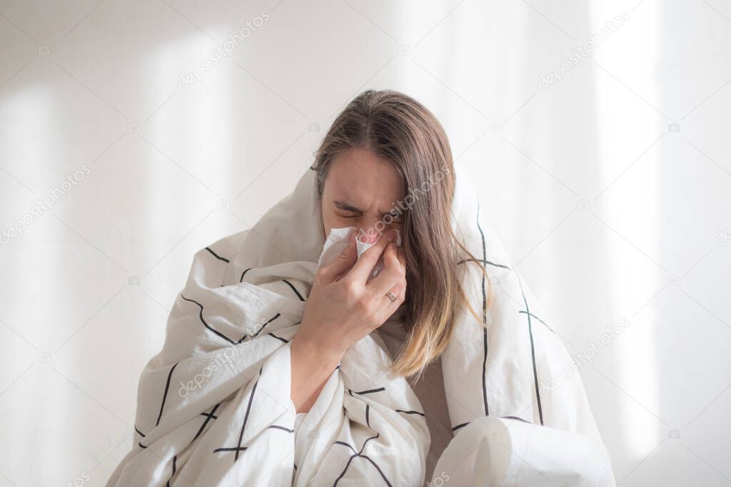 Ill woman under white blanket blowing her nose on white background. Illness treatment, home isolation, quarantine. Copy space