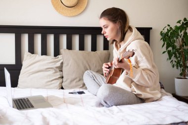 Genç ve neşeli bir kadın gitar çalmayı internetten öğreniyor. Evde yeni yetenekler öğreniyorum. Uzaklık ev eğitimi.