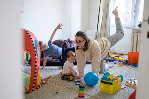 Η μητέρα γυμνάζεται σε βρώμικο παιδικό δωμάτιο με παιδιά τριγύρω. Οικογενειακός τρόπος ζωής. Αρχική προπόνηση κοινωνική αποστασιοποίηση αυτο απομόνωση — Φωτογραφία Αρχείου