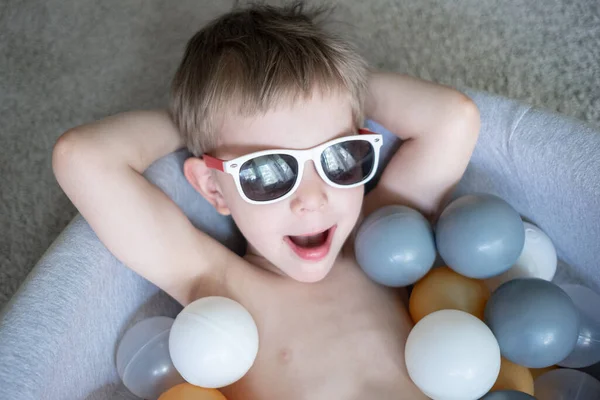 Güneş gözlüklü çocuk evde top havuzunda yüzüyor. Evde kendini izole etme karantina eğlencesi. Evde tatil turizmi — Stok fotoğraf