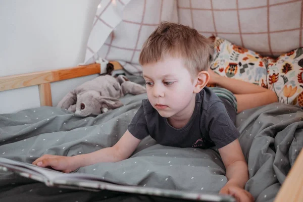 Chłopak z przedszkola czyta książkę leżącą w jego łóżku. spędzać czas w domu. Koncepcja rodziny stylu życia — Zdjęcie stockowe