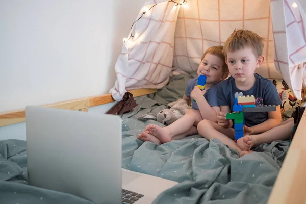 Chłopcy siedzą na łóżku z laptopem. Oglądanie kreskówek, wideo czat, edukacja na odległość. Zamknięcie, kwarantanna. Rodzina stylu życia — Zdjęcie stockowe