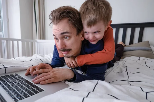Człowiek próbujący pracować przez laptopa w domu podczas kwarantanny z małym chłopcem skaczącym na łóżku. Życie rodzinne podczas samoizolacji. Zostań w domu. Praca w domu, domowe biuro — Zdjęcie stockowe