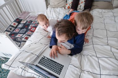 Karantina sırasında evde dizüstü bilgisayarla çalışmaya çalışan bir çocuk yatağa atlıyor. Kişisel izolasyon sırasında aile hayatı. Evde kal. Evden çalış, ev ofisinden.