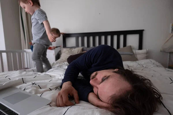 Hombre tratando de trabajar a través de la computadora portátil en casa durante la cuarentena con el niño saltando en la cama. Vida familiar durante el aislamiento personal. Quédate en casa. Trabajo desde casa, oficina en casa —  Fotos de Stock