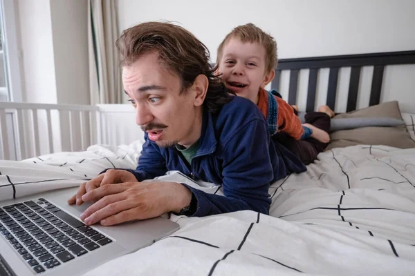 Człowiek próbujący pracować przez laptopa w domu podczas kwarantanny z małym chłopcem skaczącym na łóżku. Życie rodzinne podczas samoizolacji. Zostań w domu. Praca w domu, domowe biuro — Zdjęcie stockowe