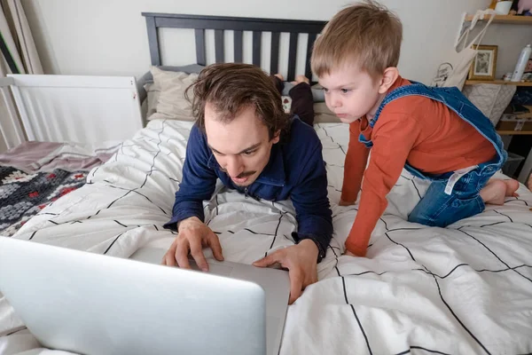 Człowiek próbujący pracować przez laptopa w domu podczas kwarantanny z małym chłopcem skaczącym na łóżku. Życie rodzinne podczas samoizolacji. Zostań w domu. Praca w domu, domowe biuro — Zdjęcie stockowe