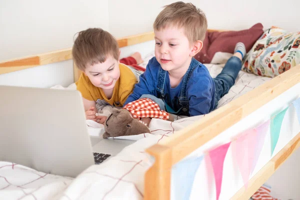 Dwóch chłopców leżących w domu na łóżku i patrzących na laptopa, robiących czat wideo, edukację na odległość, uczących się nowych, oglądających kreskówki. — Zdjęcie stockowe