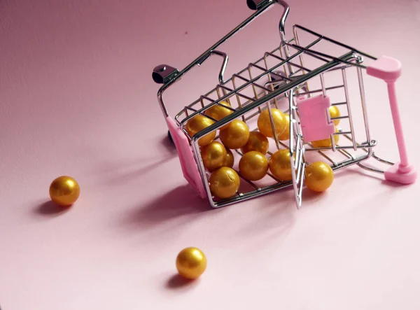 购物车。在粉红色的背景下, 下跌的超市手推车满是金色的球。消费主义概念照片. — 图库照片