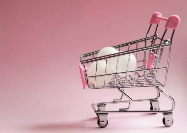 购物车。超市手推车上全是白色的球在粉红色的背景。消费主义概念照片. — 图库照片