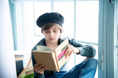 Çocuk portresi okuyor ve pencere kenarında oturuyor. 