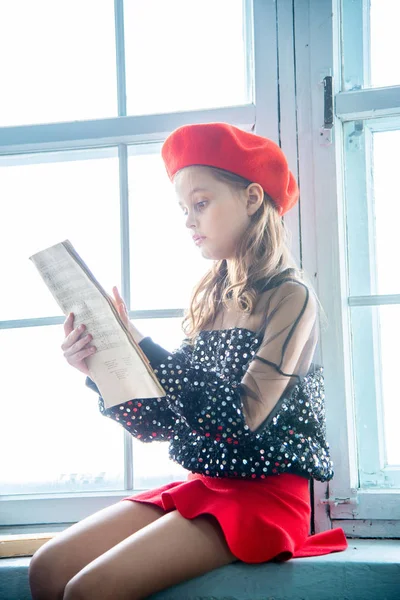 穿着红色贝雷帽和闪闪发光的毛衣的女孩 阅读音乐笔记本 坐在窗台上 — 图库照片