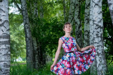 Genç ve güzel bir kadın huş ağaçlarının yanında poz veriyor. Çiçekli elbiseler giyiyor.