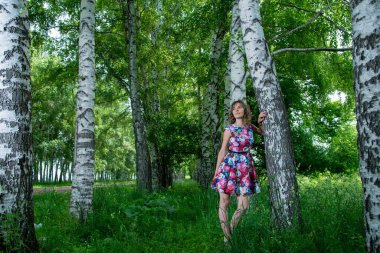 Genç ve güzel bir kadın huş ağaçlarının yanında poz veriyor. Çiçekli elbiseler giyiyor.