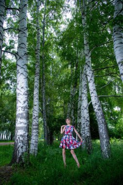 Genç ve güzel bir kadın huş ağaçlarının yanında poz veriyor. Çiçekli elbiseler giyiyor.