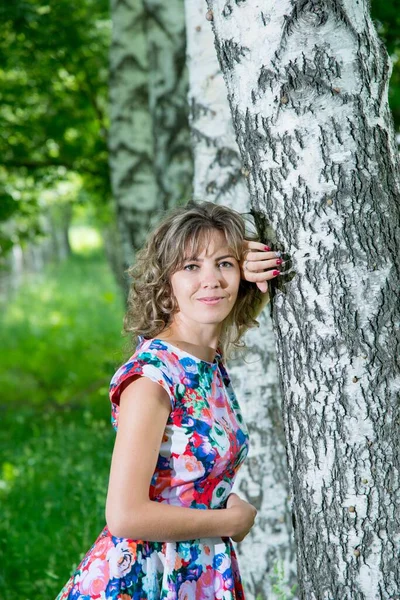 花のドレスを着た白樺の木の近くにポーズをとる若い美しい女性 — ストック写真
