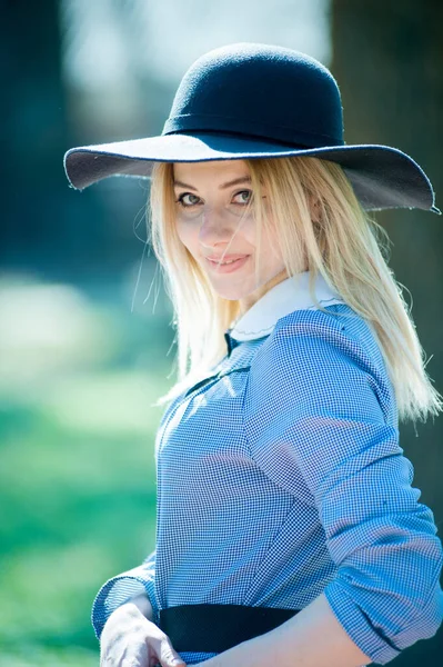 若いです可愛いですブロンド女性で青いドレスと黒の帽子を身に着けています公園で晴れた日の背景に — ストック写真