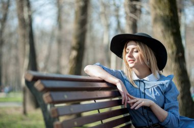 Park arka planında bankta oturan sarışın genç bir kadının portresi.