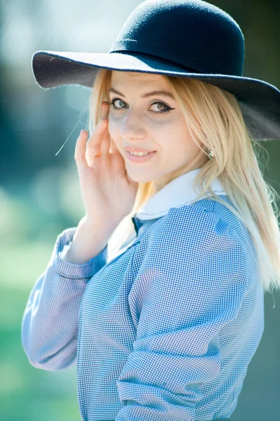 若いです可愛いですブロンド女性で青いドレスと黒の帽子を身に着けています公園で晴れた日の背景に — ストック写真