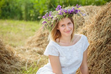 Genç güzel sarışın kız beyaz elbise ve kır çiçekleri haystack oturan bir çelenk. Yaz Köyü