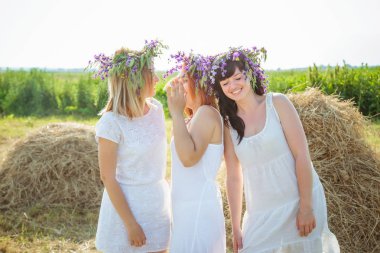 Üç genç güzel kız beyaz elbiseler ve kır çiçekleri stayingng Samanlık ve gülerek yakınındaki çelenk. Yaz Köyü