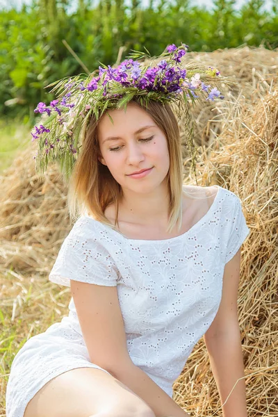Giovane bella ragazza bionda in un abito bianco e una corona di fiori selvatici seduti vicino pagliaio. Estate nel villaggio — Foto Stock