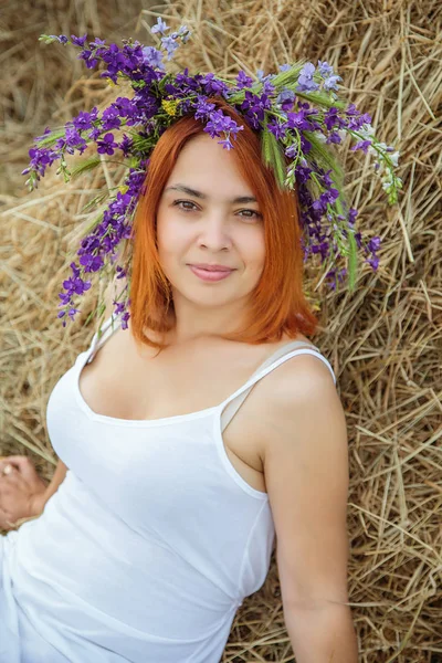 Giovane bella ragazza con i capelli rossi in un abito bianco e una corona di fiori selvatici seduti vicino pagliaio. Estate nel villaggio — Foto Stock