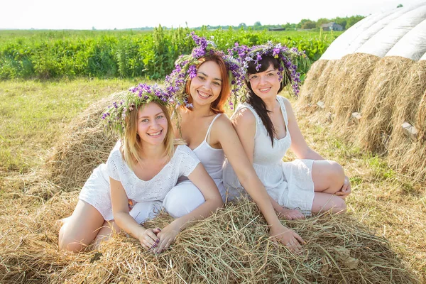 Три молодые красивые девушки в белых платьях и венках из диких цветов, сидящих возле стога сена. Лето в деревне — стоковое фото